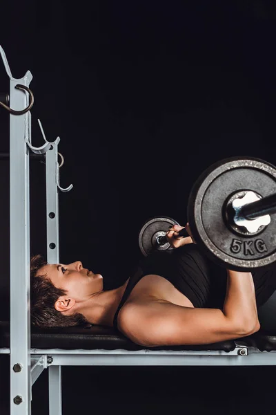 Vista laterale di giovane atleta donna che fa esercizio con bilanciere, sfondo nero — Foto stock