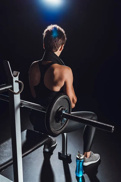 Vista trasera de la deportista con toalla sobre el cuello descansando cerca de la barra en el gimnasio con botella de agua en el suelo, fondo negro - foto de stock