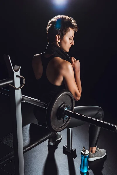 Joven deportista con toalla sobre el cuello descansando cerca de la barra en el gimnasio, fondo negro - foto de stock