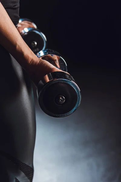 Partial view of sportswoman working out with dumbbells, black background — Stock Photo