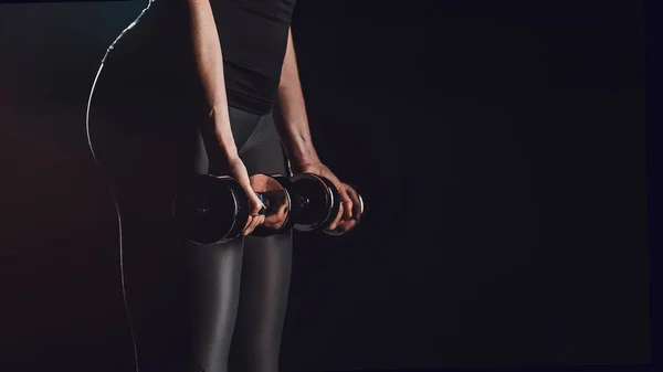 Partial view of female athlete exercising with dumbbells at gym, black background — Stock Photo
