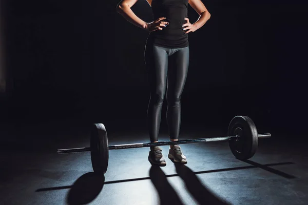 Immagine ritagliata di atleta donna in piedi con le mani in vita vicino al bilanciere sul pavimento in palestra, sfondo nero — Foto stock