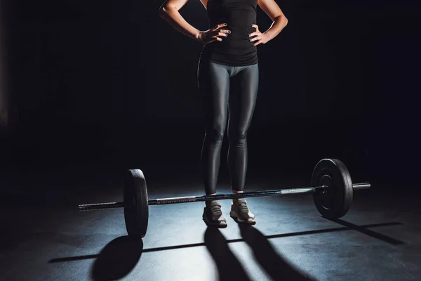 Cropped image of female athlete standing with hands on waist near barbell on floor at gym, black background — Stock Photo