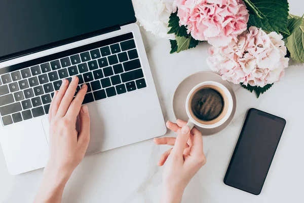 Vista cortada da mulher usando laptop e segurando xícara de café na mesa com flores de smartphone e hortensia — Fotografia de Stock