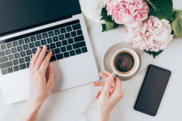 Cropped view of woman using laptop and holding cup of coffee on tabletop with smartphone and hortensia flowers — Stock Photo