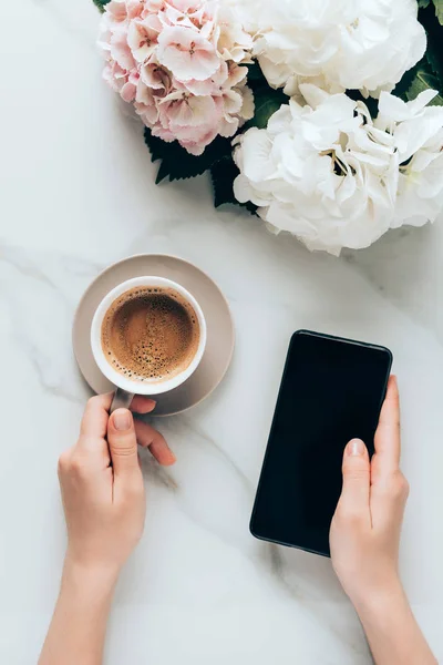 Vista cortada da mulher com smartphone e xícara de café na superfície de mármore com flores hortensia — Fotografia de Stock