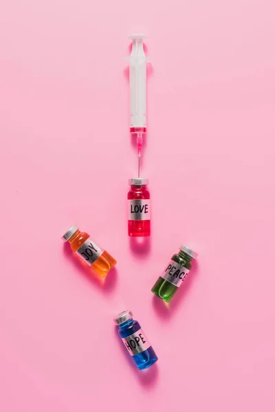 Top view of syringe and bottles with love, hope, joy and peace vaccine signs on pink tabletop — Stock Photo