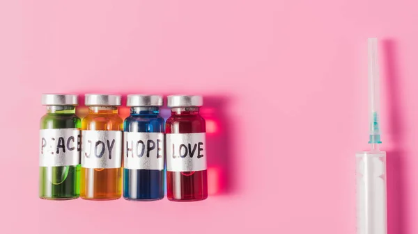 Вид зверху шприц і пляшки з любов'ю, надією, радістю і миром ознаки вакцини в ряд на рожевій стільниці — стокове фото