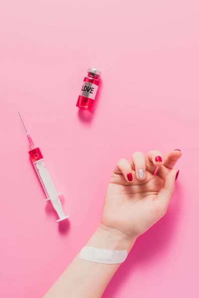 Inyección recortada de la mano de las mujeres acostadas en la superficie de color rosa con jeringa y frasco con signo de la vacuna del amor - foto de stock