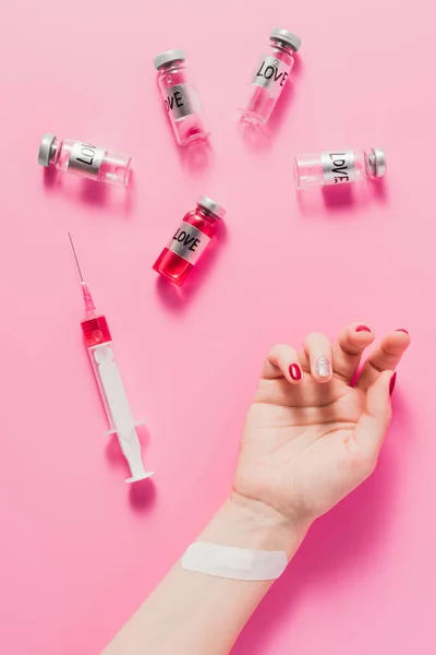 Inyección recortada de la mano de las mujeres acostadas en la superficie de color rosa con jeringa y frascos con signos de amor vacuna - foto de stock