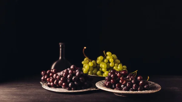 Frische reife Trauben auf Vintage-Tellern und eine Flasche Wein auf einem Holztisch auf schwarz — Stockfoto