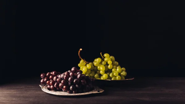 Vista de cerca de uvas rojas y blancas maduras frescas en placas vintage sobre mesa de madera sobre negro - foto de stock