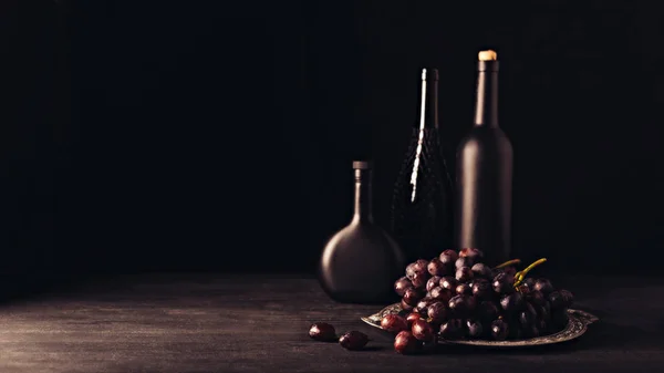 Uvas rojas maduras frescas en plato vintage y botellas de vino sobre mesa de madera sobre negro - foto de stock