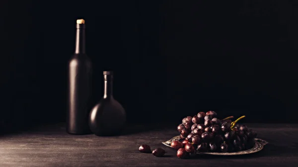 Uvas maduras frescas em prato vintage e garrafas de vinho em mesa de madeira em preto — Fotografia de Stock