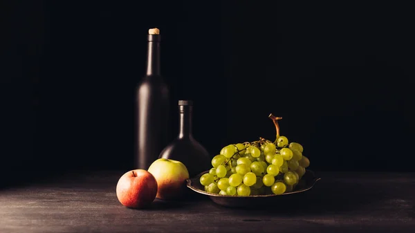 Uvas frescas maduras en plato, manzanas y botellas de vino sobre mesa de madera sobre negro - foto de stock