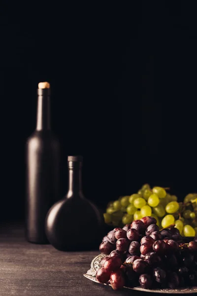 Uve fresche mature rosse e bianche e bottiglie di vino su tavolo di legno su nero — Foto stock