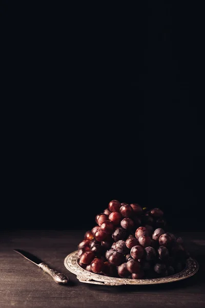 Close-up view of fresh ripe red grapes on vintage plate and knife on wooden table on black — Stock Photo