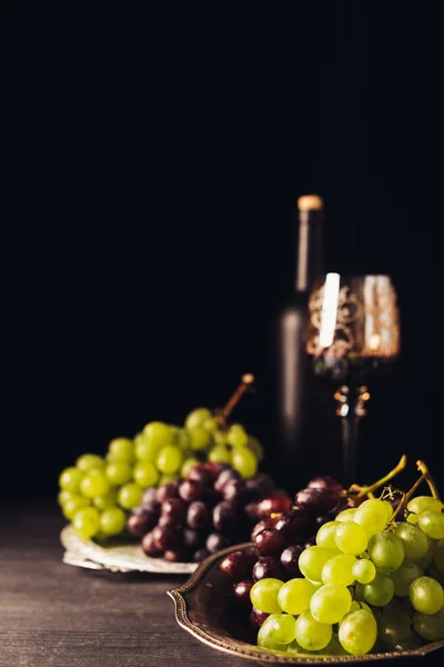 Uvas rojas y blancas sobre platos antiguos y copa con botella de vino sobre mesa de madera sobre negro - foto de stock