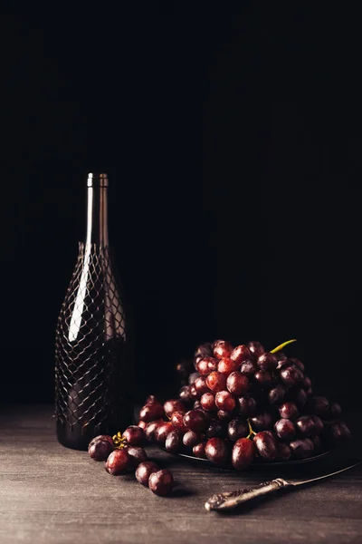 Uvas vermelhas maduras frescas em prato, faca vintage e garrafa de vinho na mesa de madeira em preto — Fotografia de Stock