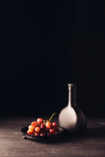 Uvas rojas maduras frescas y botella de vino sobre mesa de madera sobre negro - foto de stock