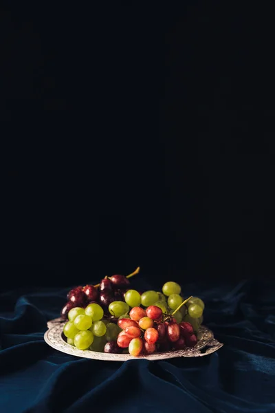 Close-up view of various types of grapes on vintage plate on dark fabric on black background — Stock Photo