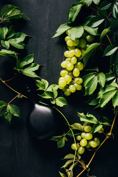 Vista superior de uvas blancas frescas con hojas verdes y botella de vino en la superficie de madera oscura - foto de stock