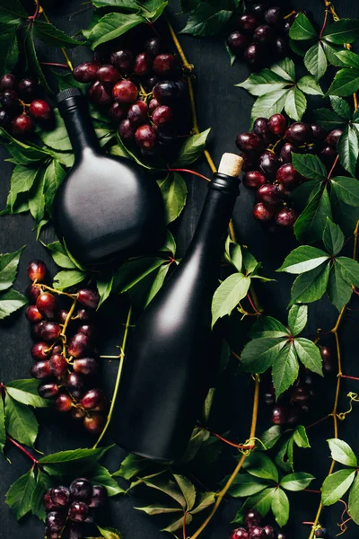 Vue de dessus des raisins frais mûrs, des feuilles vertes et des bouteilles de vin sur une surface en bois sombre — Photo de stock