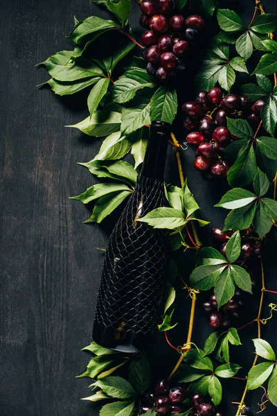 Vista superior de uvas frescas maduras, hojas verdes y botella de vino sobre fondo de madera negro - foto de stock