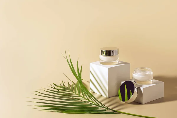 Close up view of green palm leaf, facial and body creams in glass jars on white cubes on beige background — Stock Photo