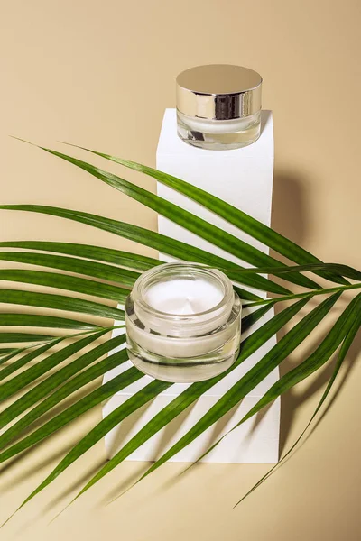 Close up view of green palm leaf and body creams in glass jars on white cubes on beige background — Stock Photo