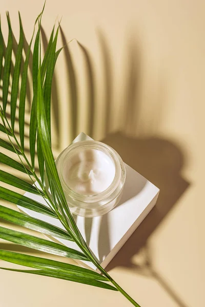 Близкий вид увлажняющего крема в стеклянной банке с пальмовым листом на бежевом фоне — стоковое фото
