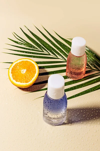 Close up view of micellar water in bottles, green palm leaf and orange half with water drops on beige backdrop — Stock Photo