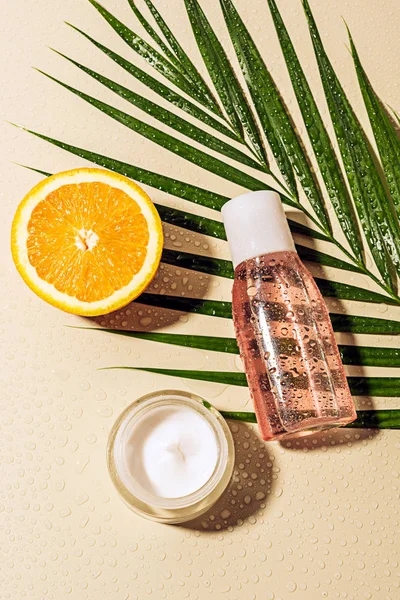 Vista dall'alto di crema viso e lozione, foglia di palma e metà arancione con gocce d'acqua su sfondo beige — Foto stock