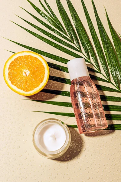 Vue du dessus de crème et lotion pour le visage, feuille de palmier et moitié orange avec des gouttes d'eau sur fond beige — Photo de stock