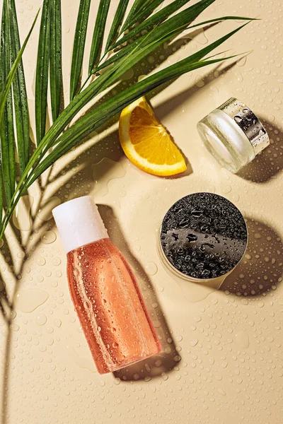 Acostado plano con productos para el cuidado de la piel, hoja de palma verde y rebanada de naranja con gotas de agua sobre fondo beige - foto de stock