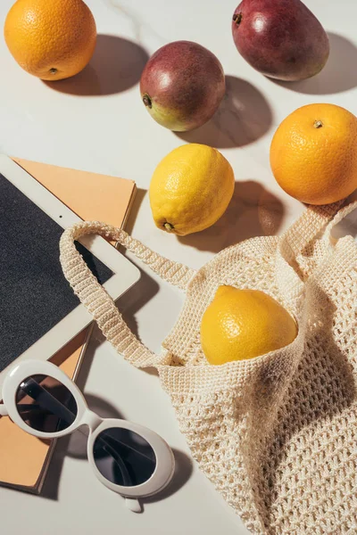 Vue rapprochée de la tablette numérique, des lunettes de soleil et du sac à ficelle avec des fruits tropicaux frais mûrs — Photo de stock