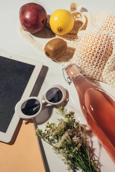 Draufsicht auf digitales Tablet, Sonnenbrille, Blumen, Glasflasche mit Sommerdrink und Saitentasche mit frischem Obst — Stockfoto