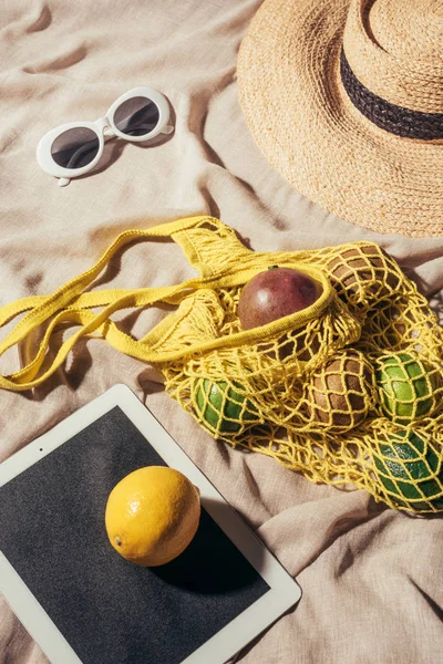 Vista de ángulo alto de la tableta digital, gafas de sol, sombrero de paja y bolsa de hilo con frutas maduras - foto de stock