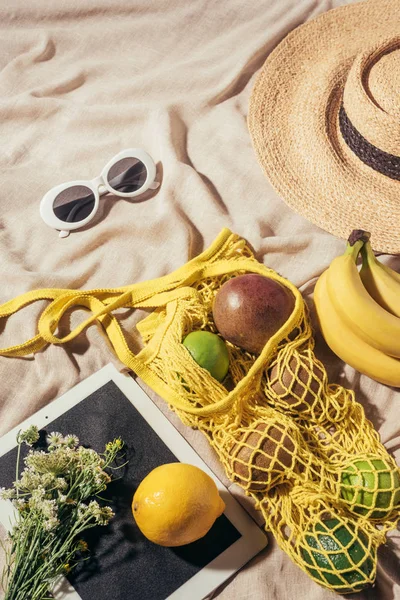 Vista superior de sombrero de paja, gafas de sol, flores, tableta digital y bolsa de cuerda con frutas maduras - foto de stock