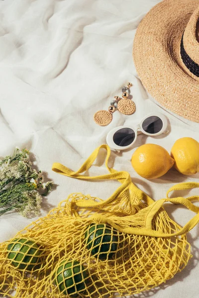 Visão de alto ângulo de chapéu de palha, óculos de sol, brincos, flores e saco de corda amarela com frutas tropicais maduras — Fotografia de Stock