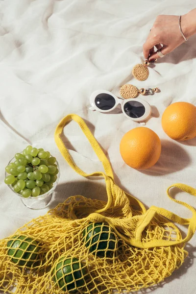 Tiro recortado de mano humana, pendientes, gafas de sol y bolsa de hilo con frutas frescas - foto de stock