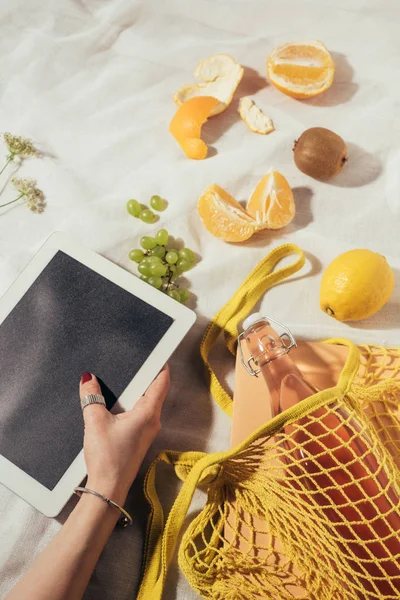 Foto recortada de la persona usando tableta digital y bolsa de cuerda con botella de vidrio y frutas frescas - foto de stock