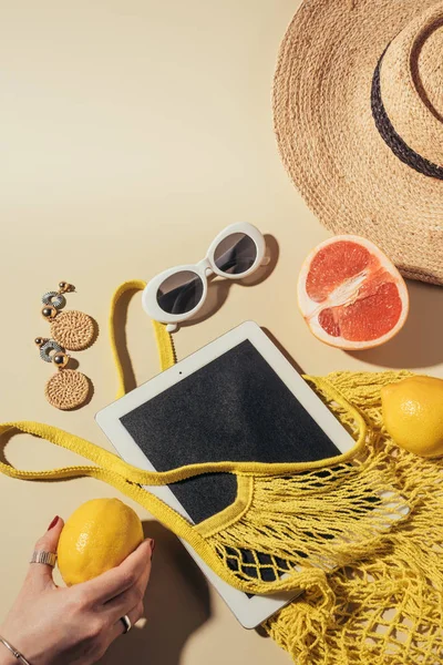 Обрезанный снимок человеческой руки с лимоном, цифровой планшет, струнный мешок и солнцезащитные очки на коричневом — стоковое фото