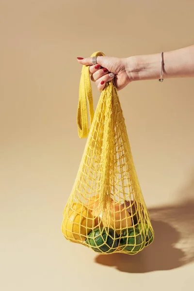 Partial view of person holding string bag with fresh tropical fruits on brown — Stock Photo