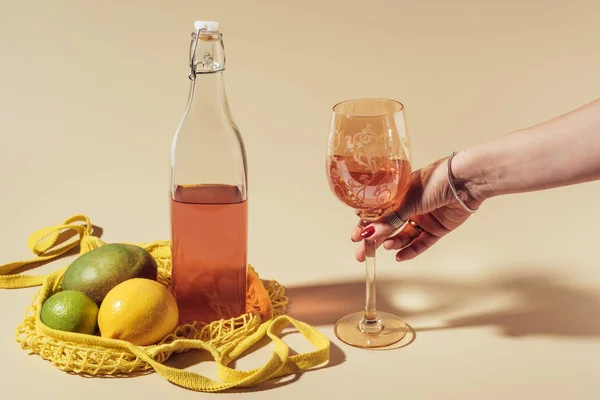 Tiro recortado de la persona sosteniendo el vidrio con la bebida, la botella y el bolso de la cuerda con frutas en marrón — Stock Photo