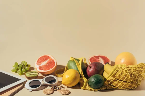 Lunettes de soleil, boucles d'oreilles, tablette numérique et fruits mûrs dans un sac à ficelle sur brun — Photo de stock