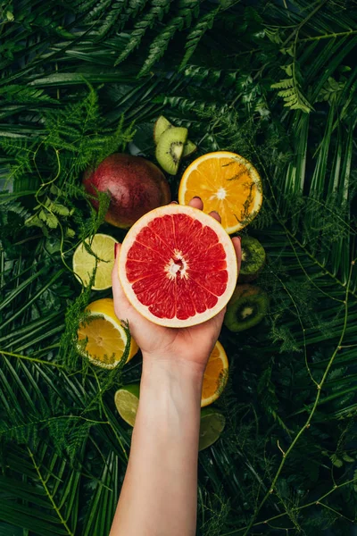 Cropped view of woman holding half of grapefruit over fruits on palm leaves — Stock Photo