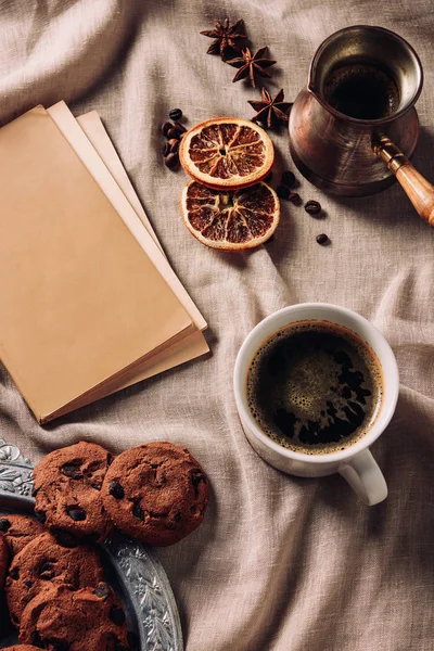 Draufsicht auf Tasse Kaffee mit Buch und Schokoladenkeksen auf beigem Tuch — Stockfoto