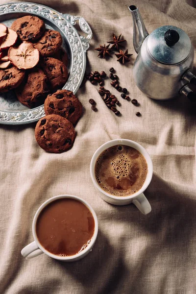 Vue du dessus des tasses de café frais avec biscuits aux pépites de chocolat et pot en métal vintage sur tissu beige — Photo de stock