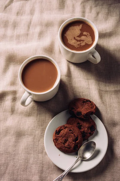 Vue du dessus des tasses de café avec biscuits aux pépites de chocolat sur plaque sur tissu beige — Photo de stock